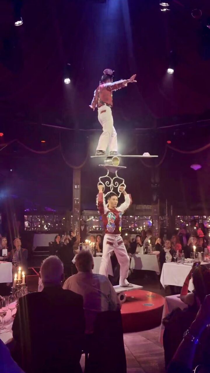One artist balancing on a board held overhead by a second artist in a mirror tent